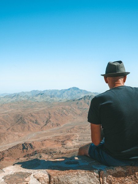 Ein Mann mit Hut sitzt auf einem Berg und schaut hinunter ins Tal und zu anderen Bergen.