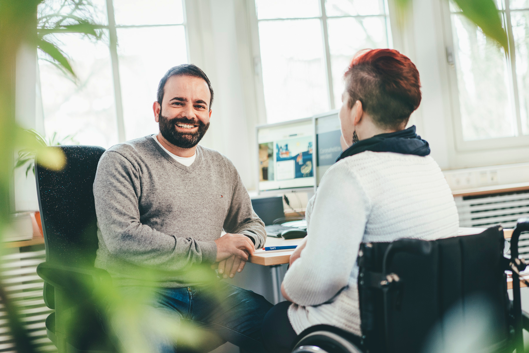 Mann sitzt lächelnd in einem Beratungsgespräch mit Rollstuhlfahrerin.