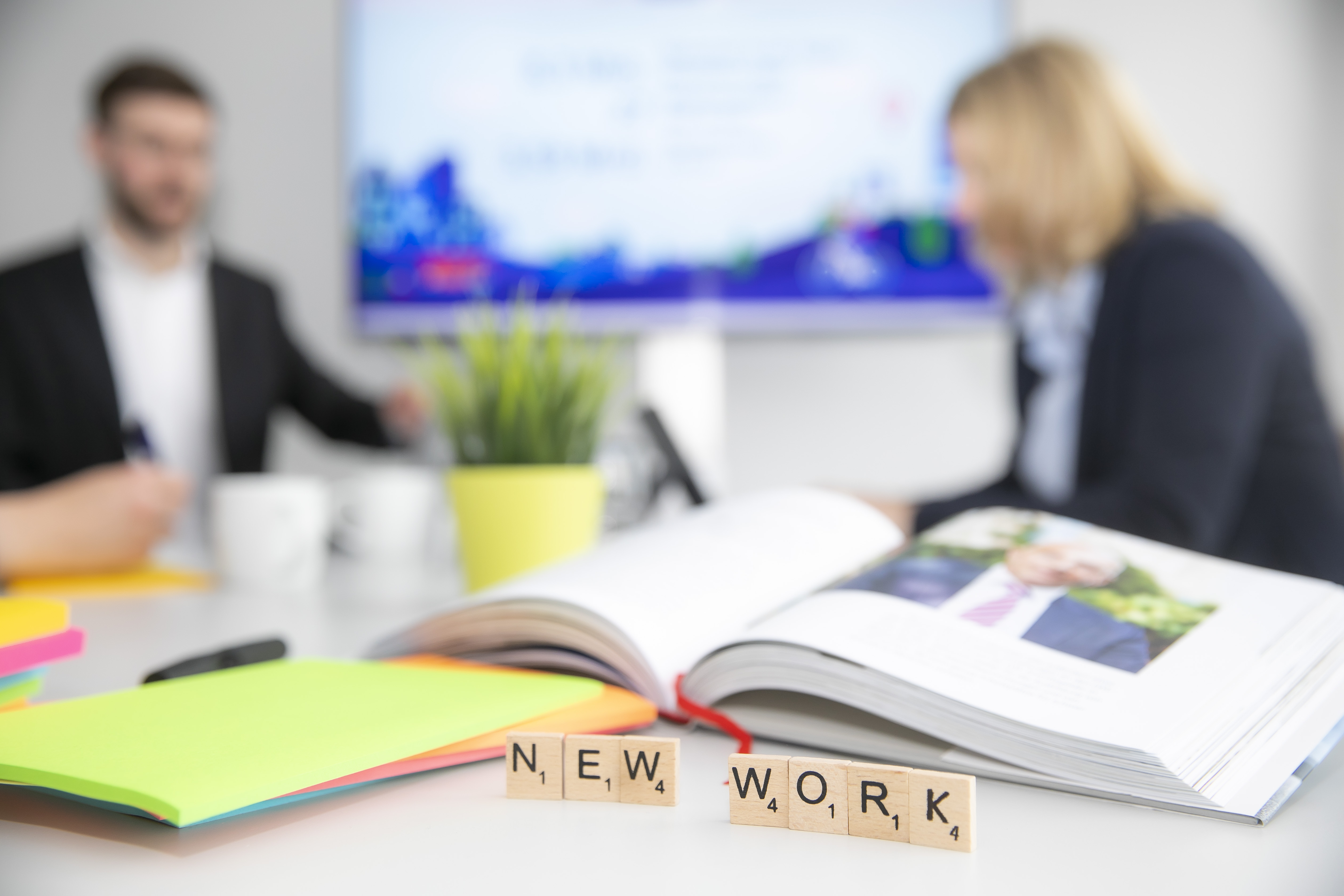 Auf einem Tisch liegt ein aufgeschlagenes Buch, daneben ein Stapel farbiger Post-Its. Im Vordergrund kleine Holzklötzchen mit dem Schriftzug New Work.