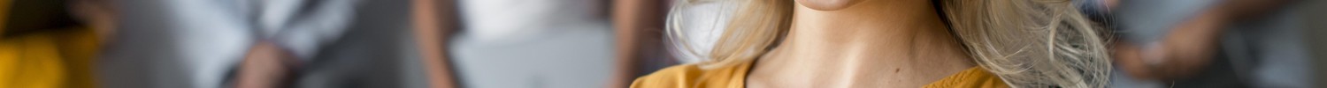Blonde Frau hält Notizbuch und lächelt freundlich in die Kamera, Kolleg:innen im Hintergrund.
