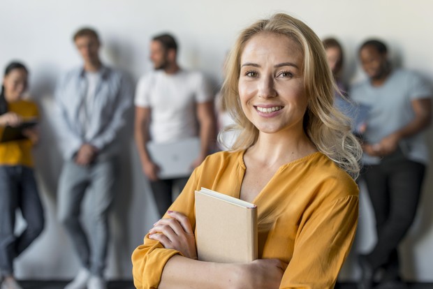 Blonde Frau mit Notizbuch lächelt in die Kamera, Kolleg:innen im Hintergrund. Blonde Frau mit Notizbuch lächelt in die Kamera, Kolleg:innen im Hintergrund.